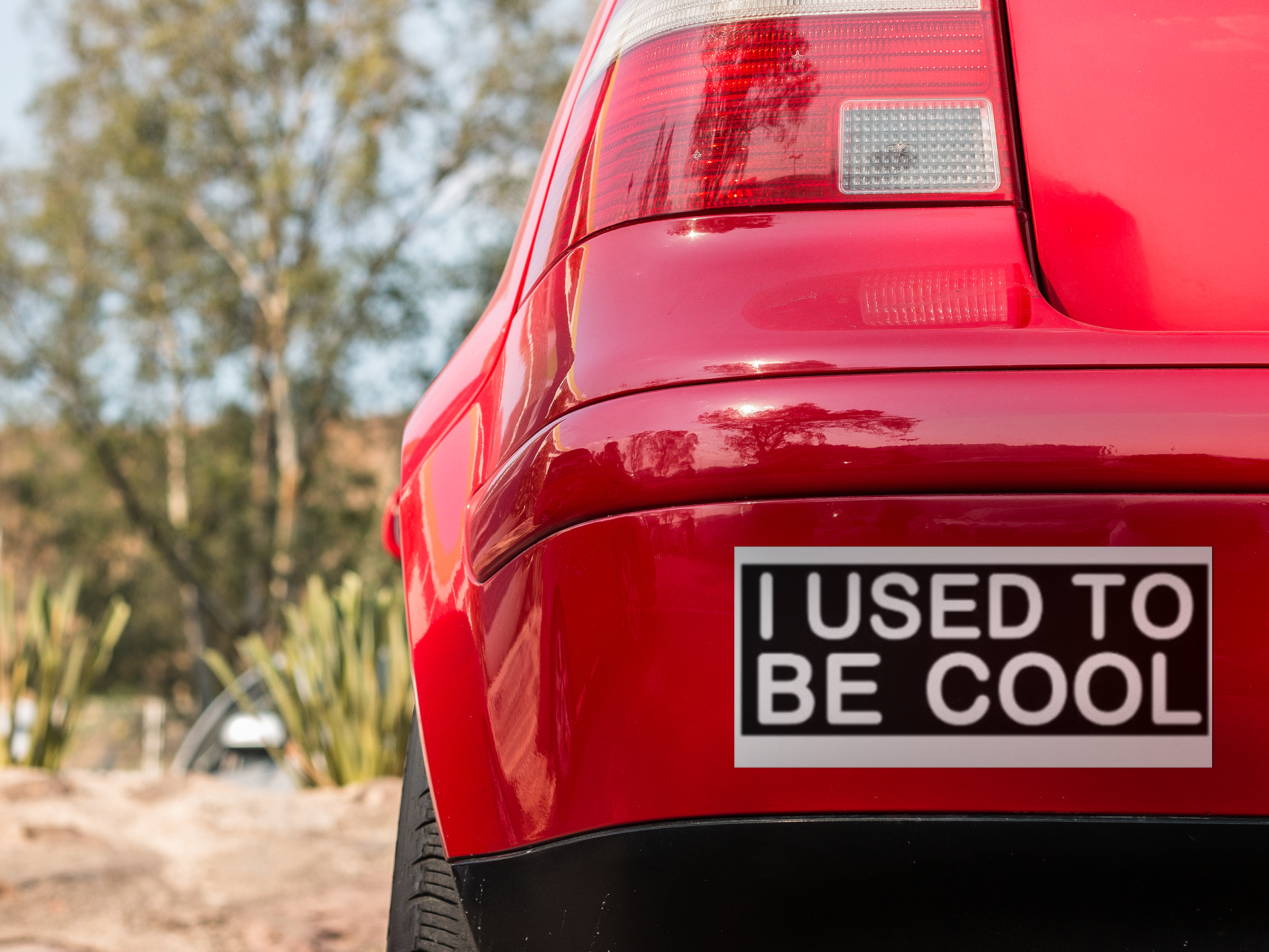 Red car with a 'I USED To Be Cool' sticker on the back, outdoors.