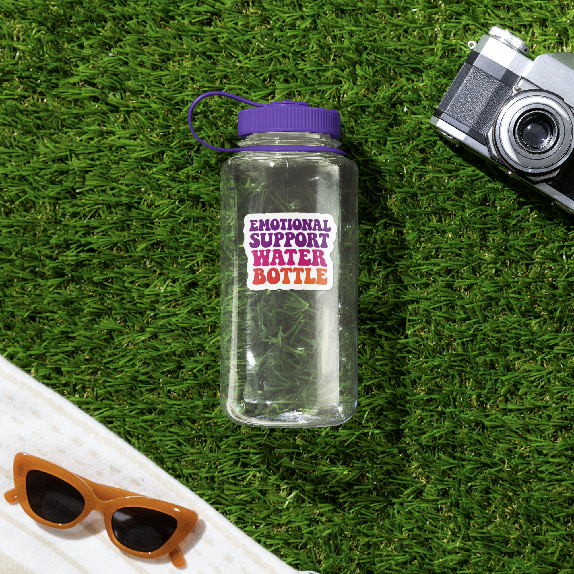 Clear water bottle with purple lid and 'Emotional Support Water Bottle' label on grass, with sunglasses and camera.