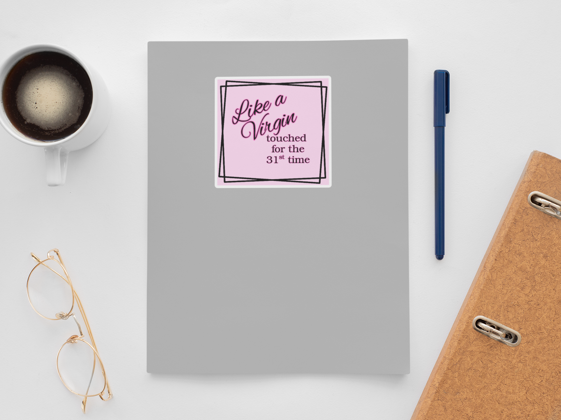 Gray notebook with a pink Like a Virgin sticker, coffee cup, glasses, pen, and phone on a white background