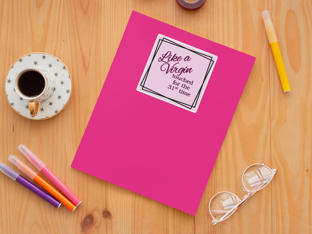 Pink notebook with a Like a Virgin sticker on a wooden surface with stationery items.