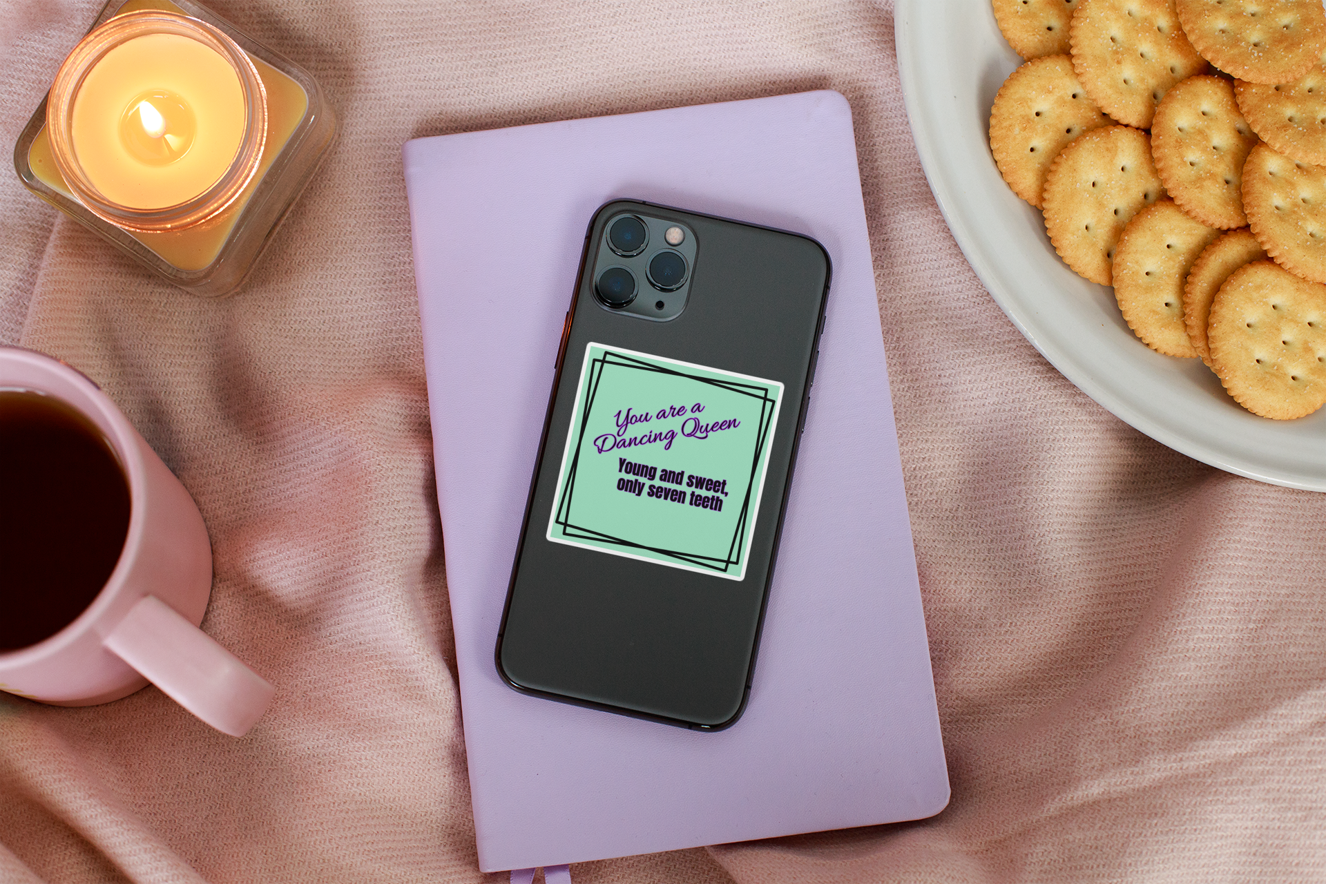 Phone with a humorous text case on a pink surface with a candle, mug, and plate of cookies.