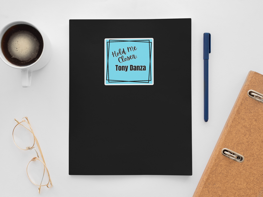 Black notebook with a blue "Hold Me Closer, Tony Danza" sticker on a white surface with a cup, pen, and glasses