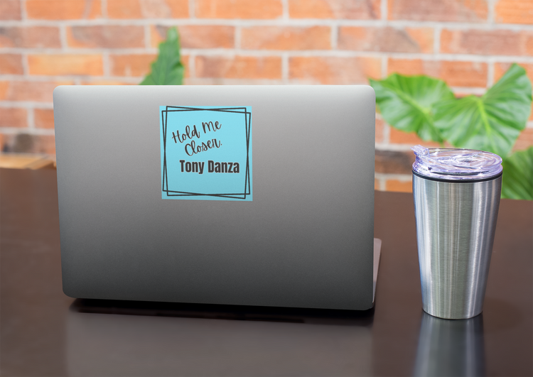 Laptop with a "Hold Me Closer, Tony Danza" on a desk with a cup, against a brick wall background