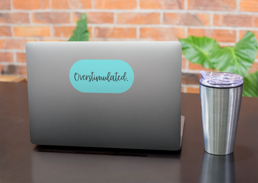 Laptop with a 'Overstimulated' sticker on a desk next to a tumbler, against a brick wall background.
