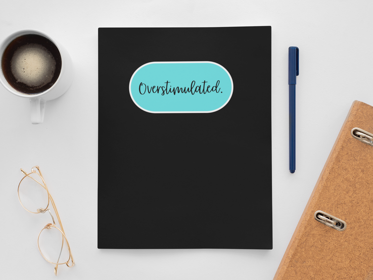 Black notebook with 'Overstimulated' sticker, coffee cup, glasses, pen, and folder on a white surface