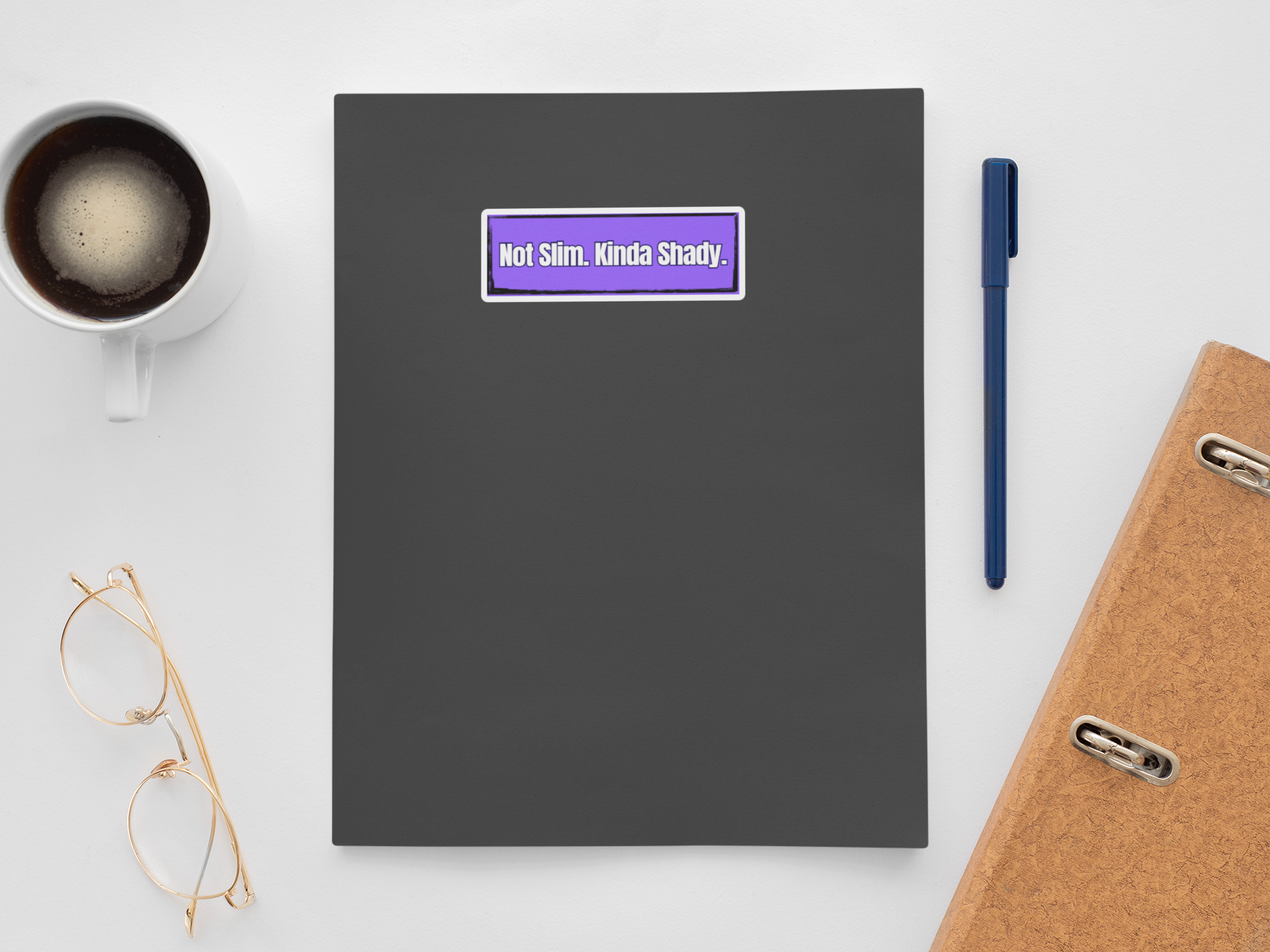Black notebook with a purple sticker, coffee cup, glasses, pen, and phone on a white surface