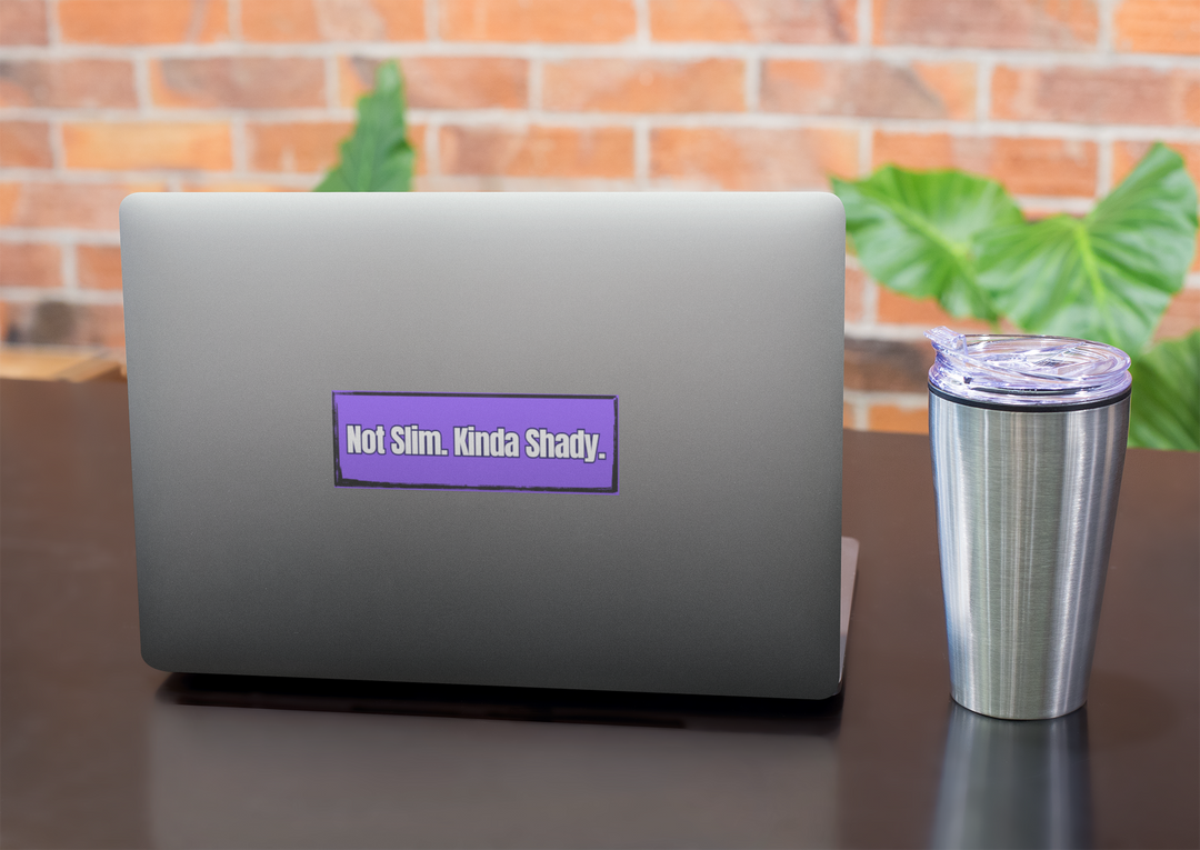 Laptop with a purple Not Slim, Kinda Shady sticker on a desk with a tumbler, against a brick wall background