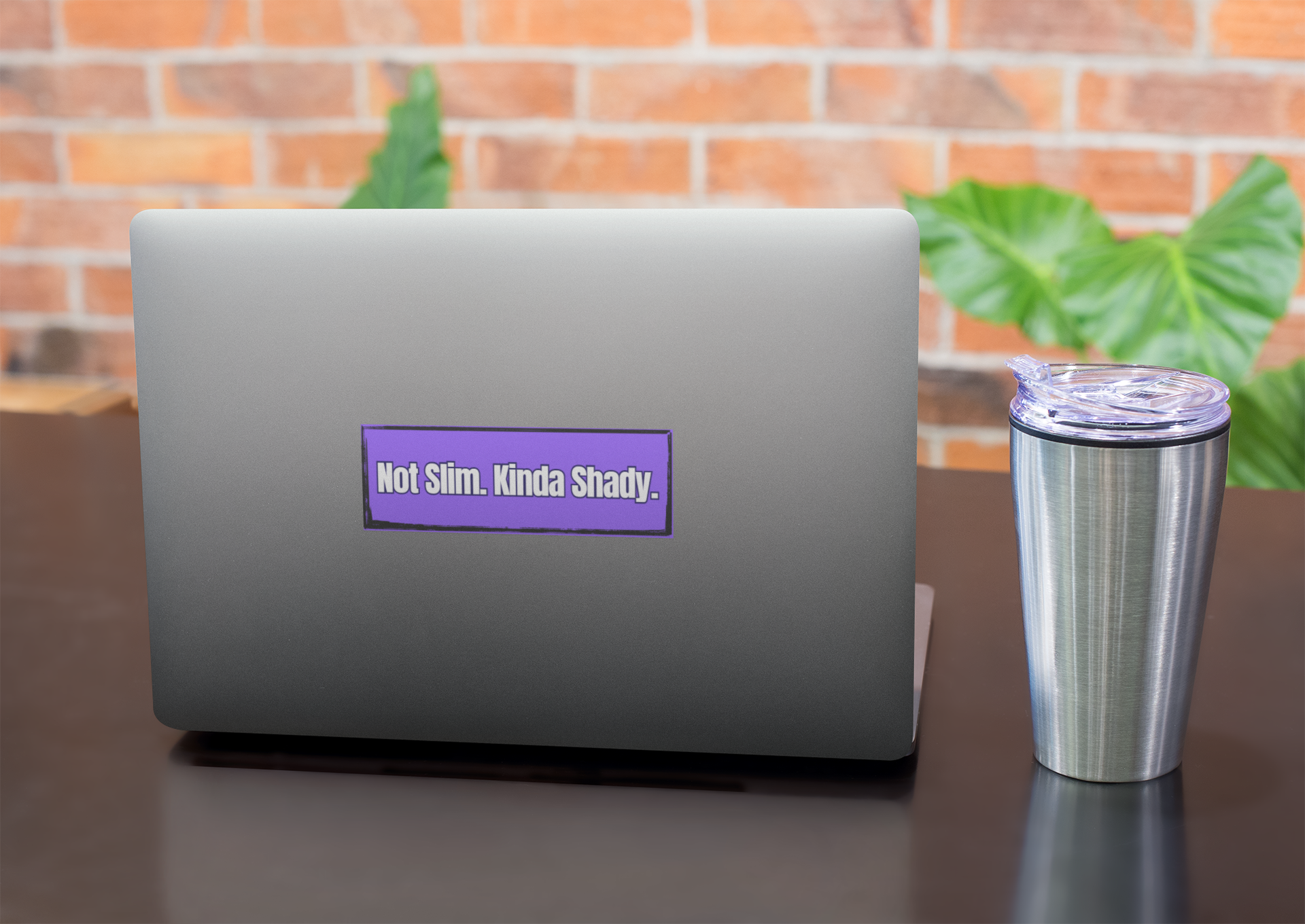 Laptop with a purple Not Slim, Kinda Shady sticker on a desk with a tumbler, against a brick wall background