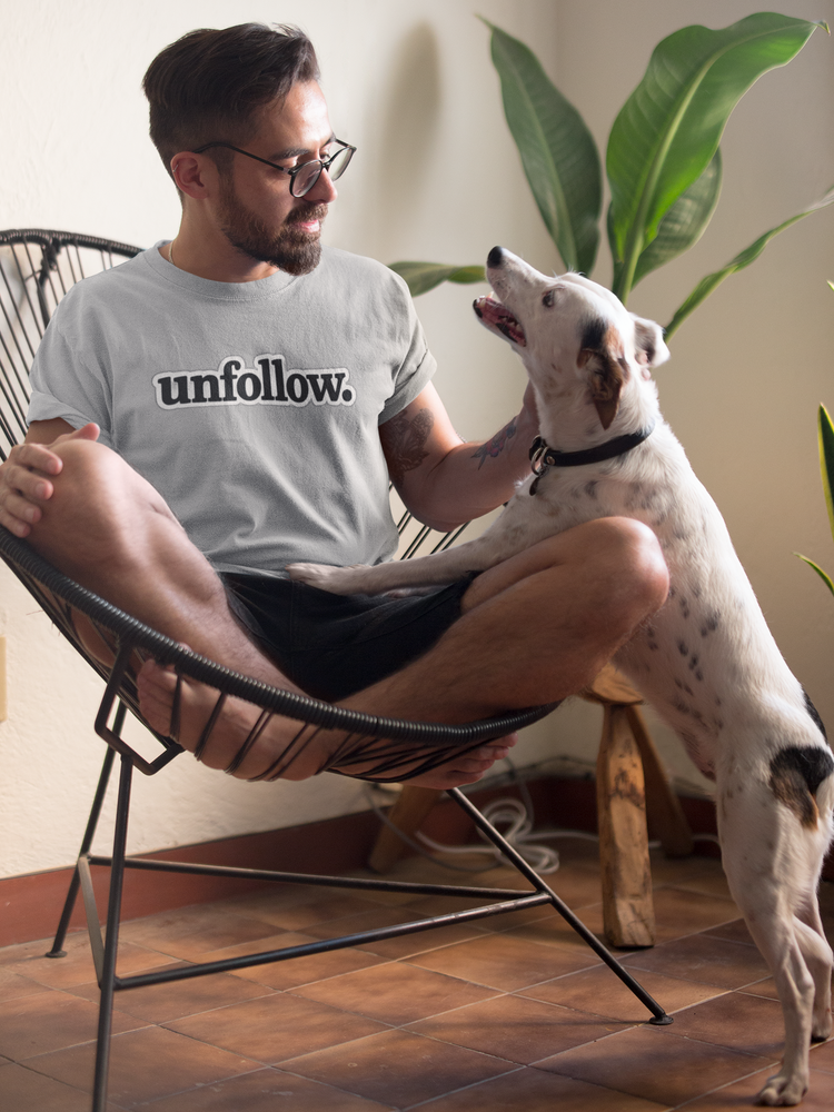 Man sitting on a chair with a dog, wearing a shirt that says 'unfollow.'