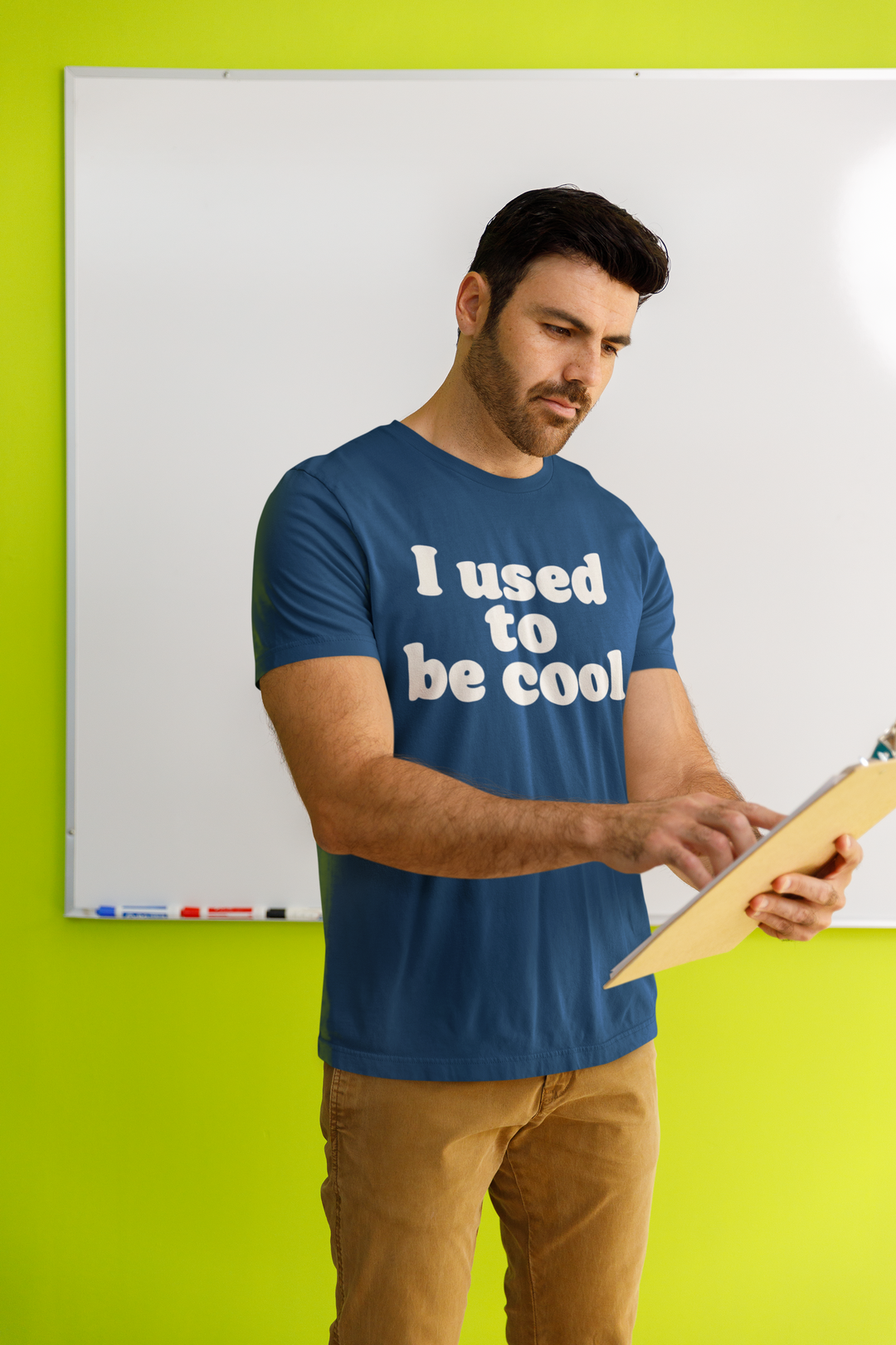 Man wearing a blue t-shirt with 'I used to be cool' text, holding a clipboard against a green wall.