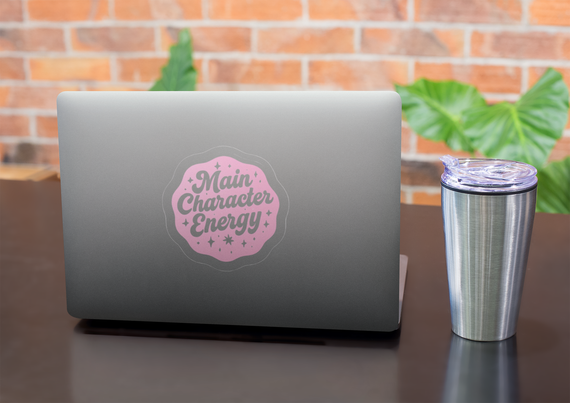 Laptop with a 'Main Character Energy' sticker on a desk with a tumbler, against a brick wall background.