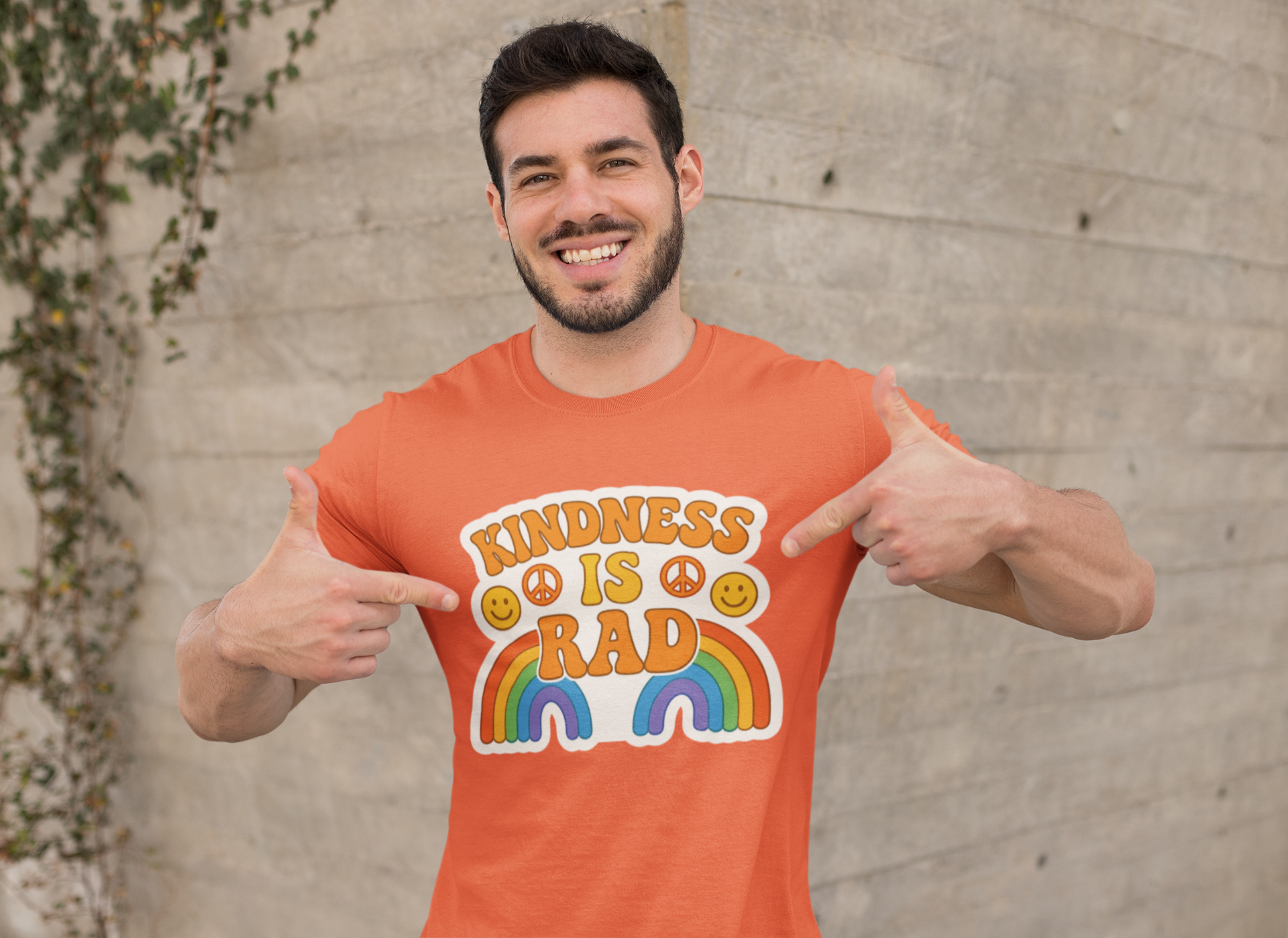 Man wearing an orange t-shirt with a rainbow with "Kindness is Rad" pointing at the shirt.