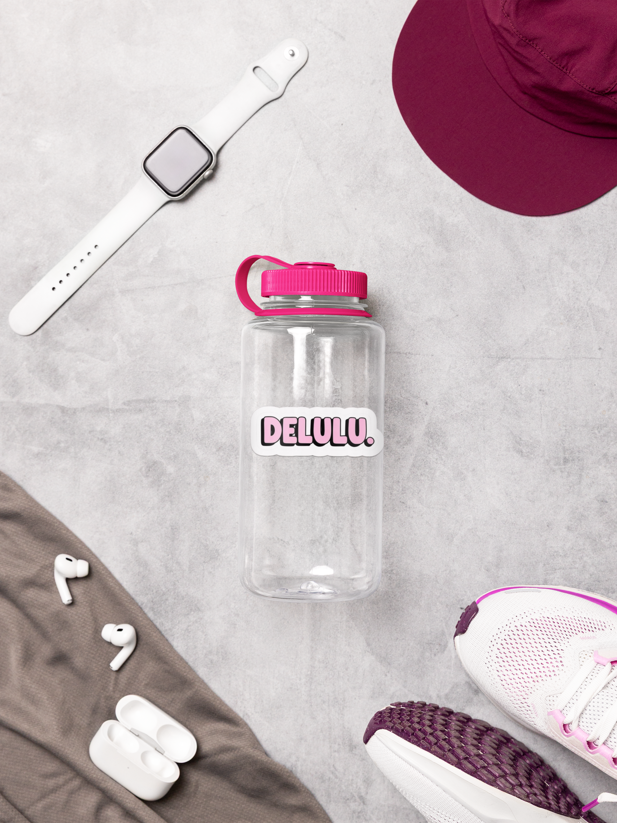 Clear water bottle with pink lid and 'DELULU' branding on a gray surface with accessories.
