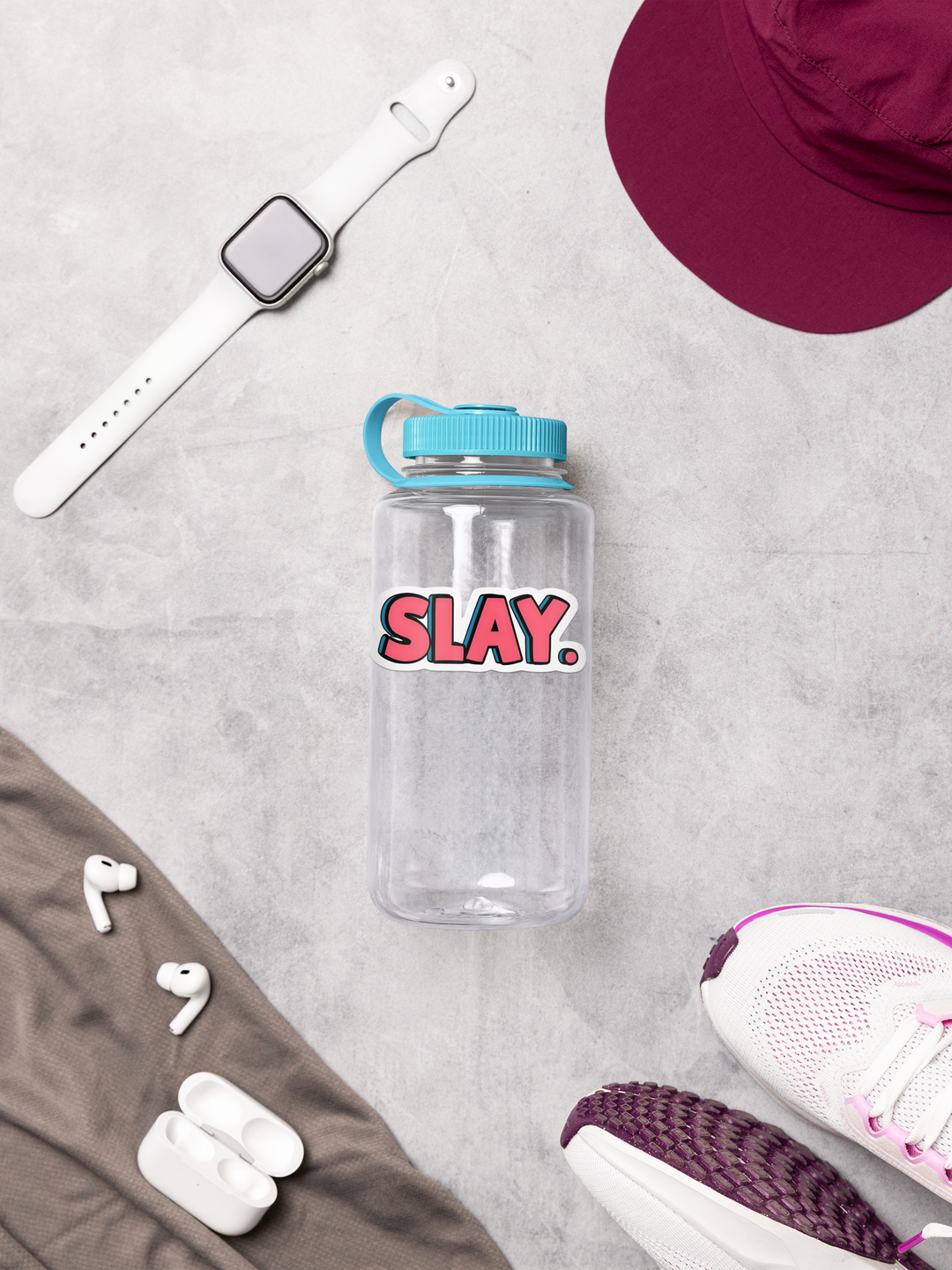 Clear water bottle with 'SLAY.' text, white sneakers, maroon hat, and white earbuds on a light gray surface.