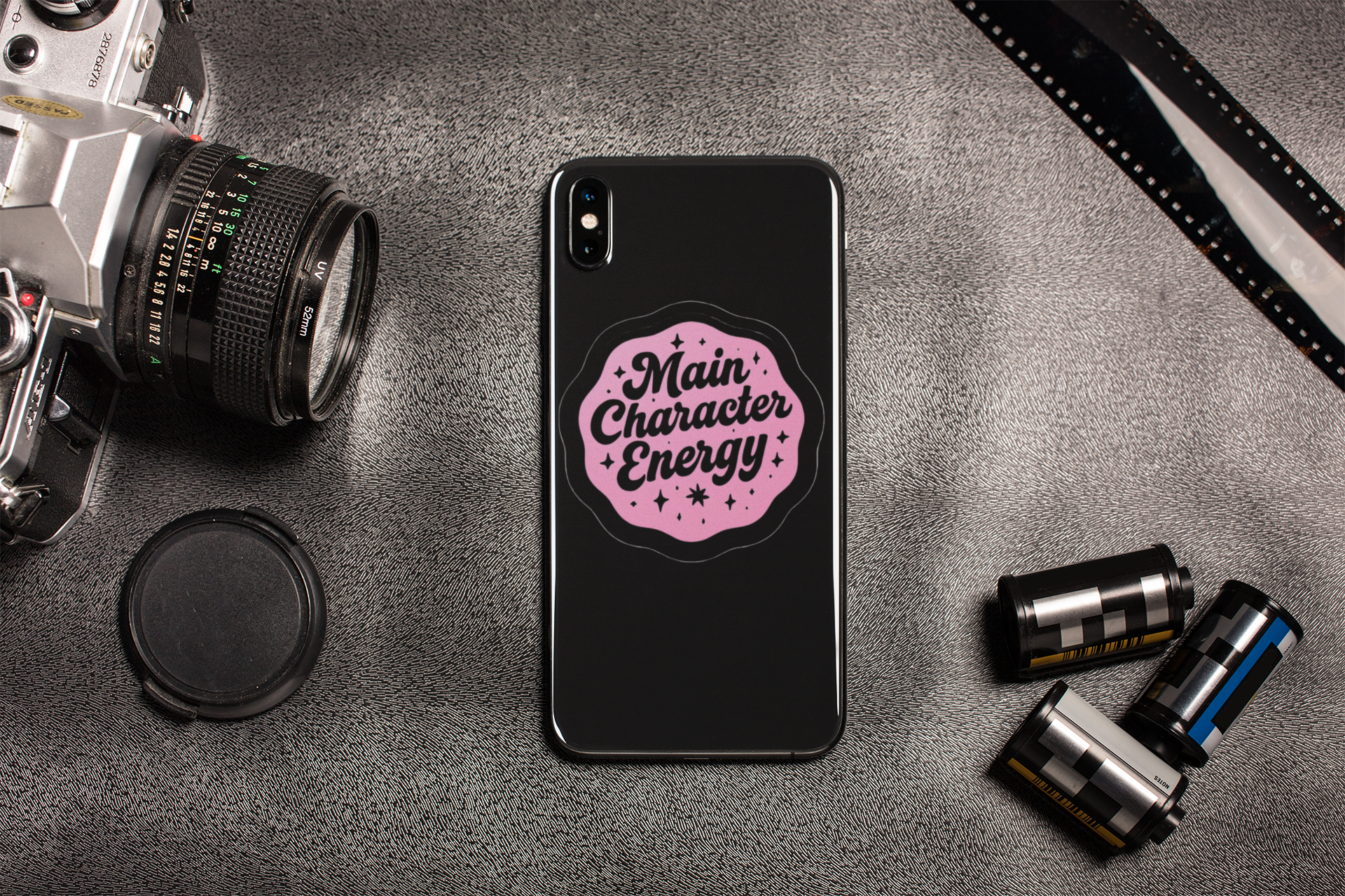 Phone case with 'Main Character Energy' design on a gray surface with camera equipment.