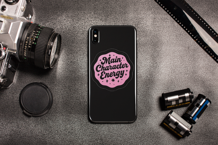 Phone case with 'Main Character Energy' design on a gray surface with camera equipment.