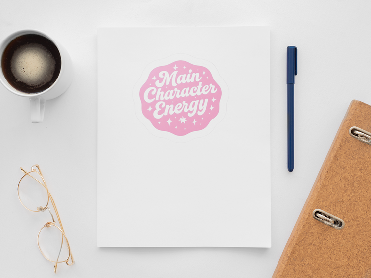 Notebook with 'Main Character Energy' sticker, coffee cup, pen, and glasses on a white surface
