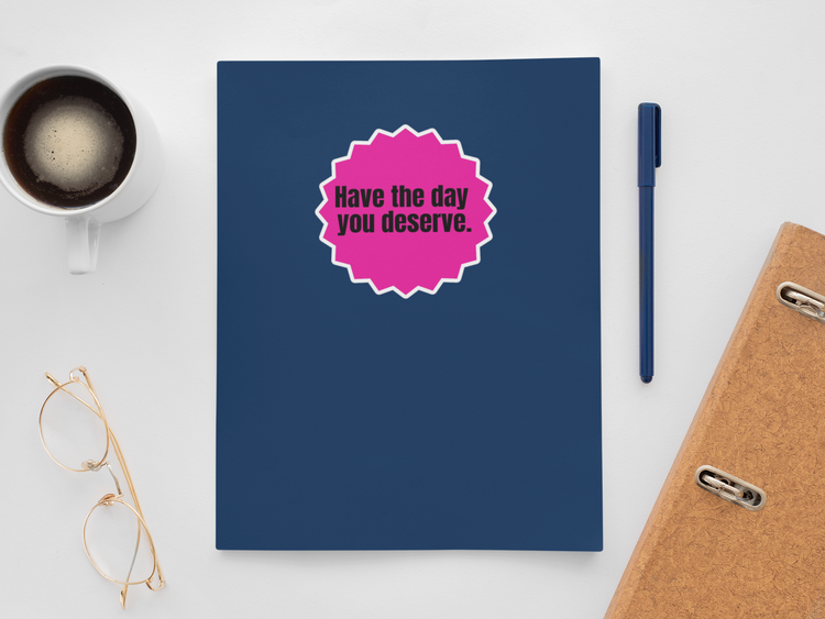 Blue notebook with a pink sticker saying 'Have the day you deserve.' on a white surface with a cup of coffee, pen, and glasses.