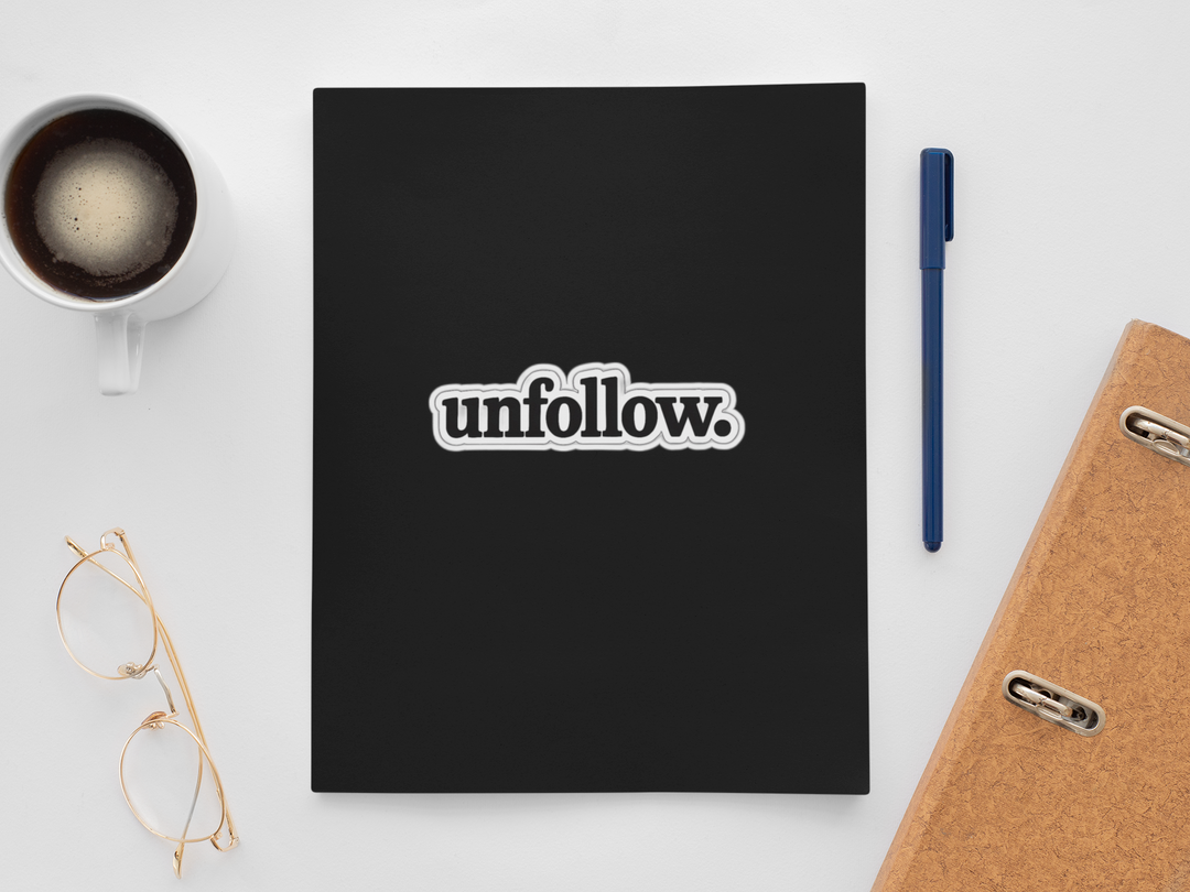 Black notebook with 'unfollow.' sticker on a white surface with a cup, pen, and glasses.
