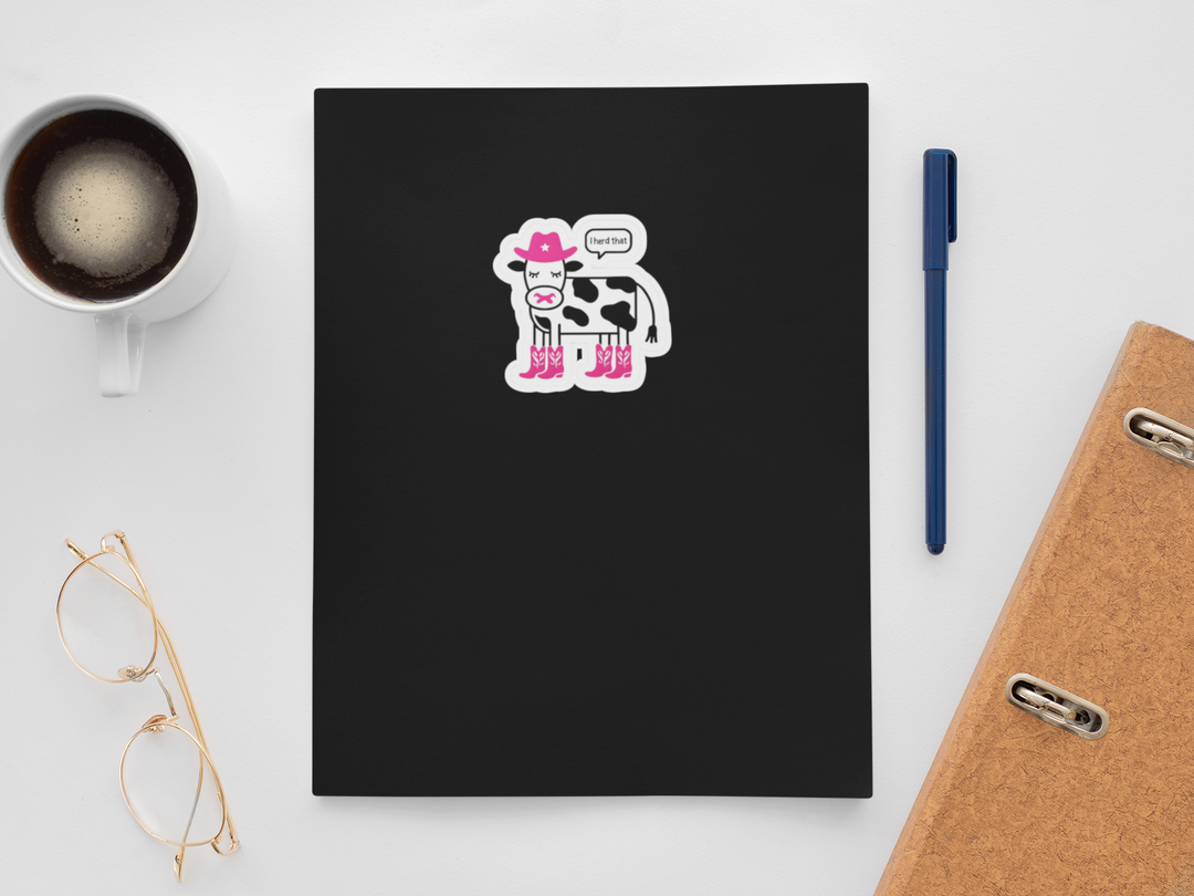 Black notebook with a cow sticker , "I herd that" on a white surface with a cup of coffee, pen, and glasses.