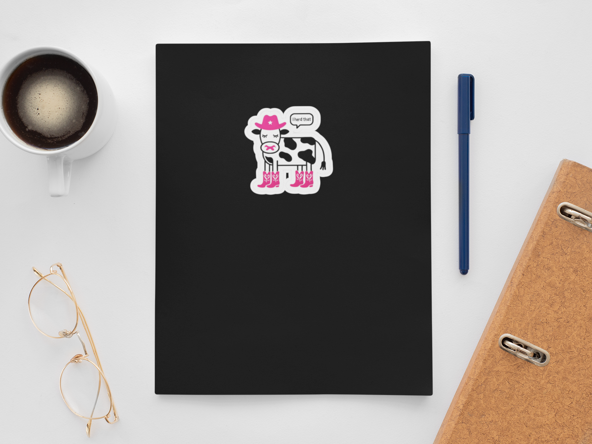 Black notebook with a cow sticker , "I herd that" on a white surface with a cup of coffee, pen, and glasses.