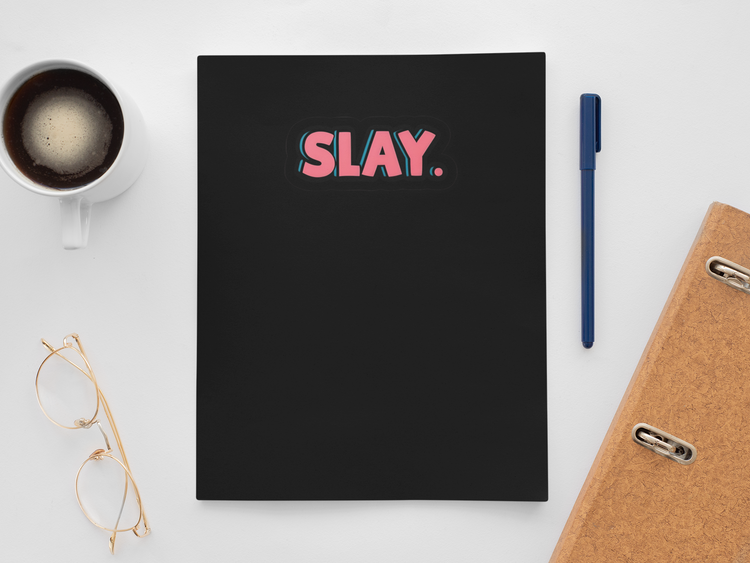 Black notebook with 'SLAY' on a white surface with a cup, pen, and glasses.