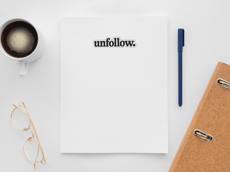 Notebook with 'unfollow.' text, coffee cup, glasses, pen, and phone on a white surface