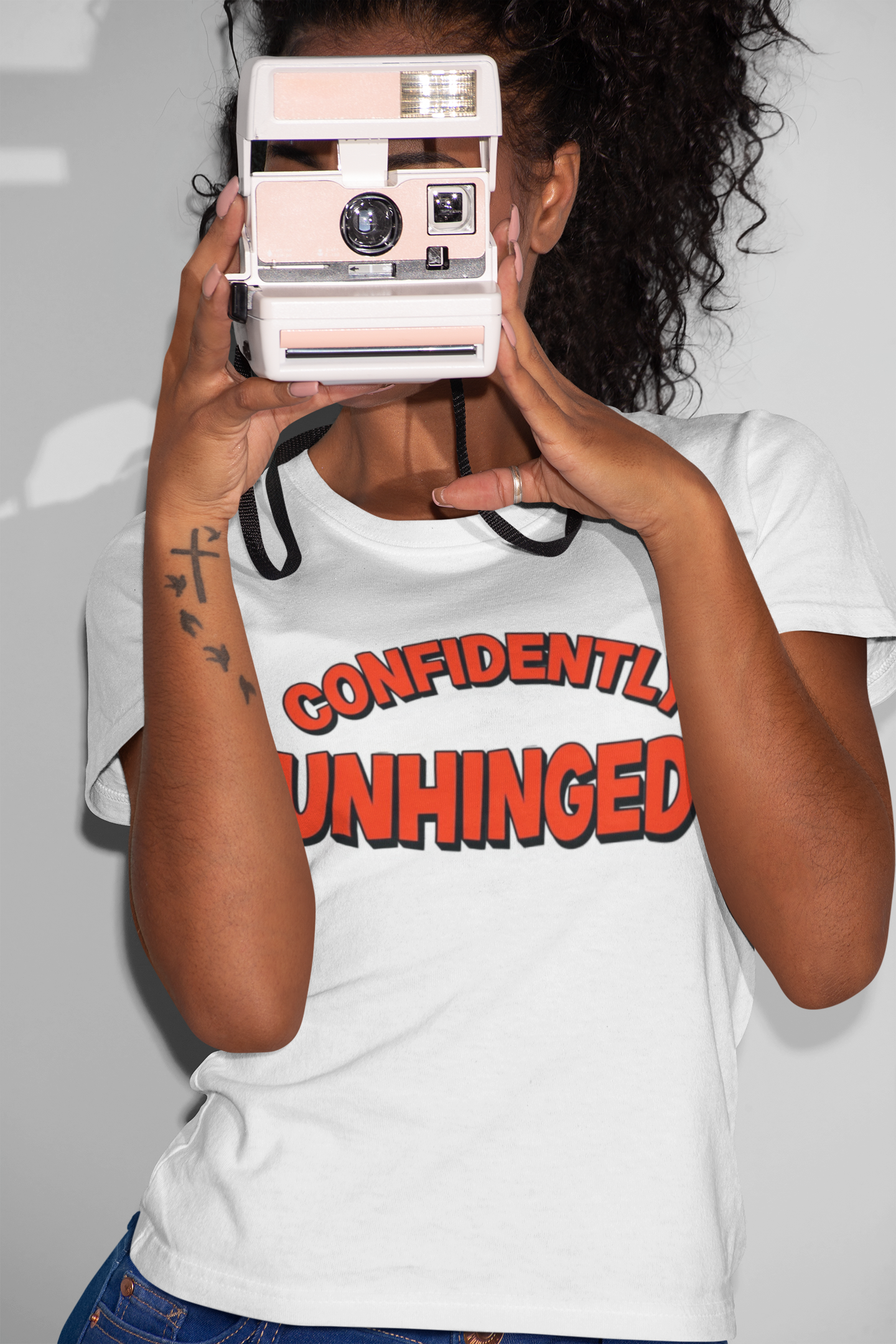 Person wearing a white t-shirt with 'CONFIDENTLY UNHINGED' text, holding a vintage camera.