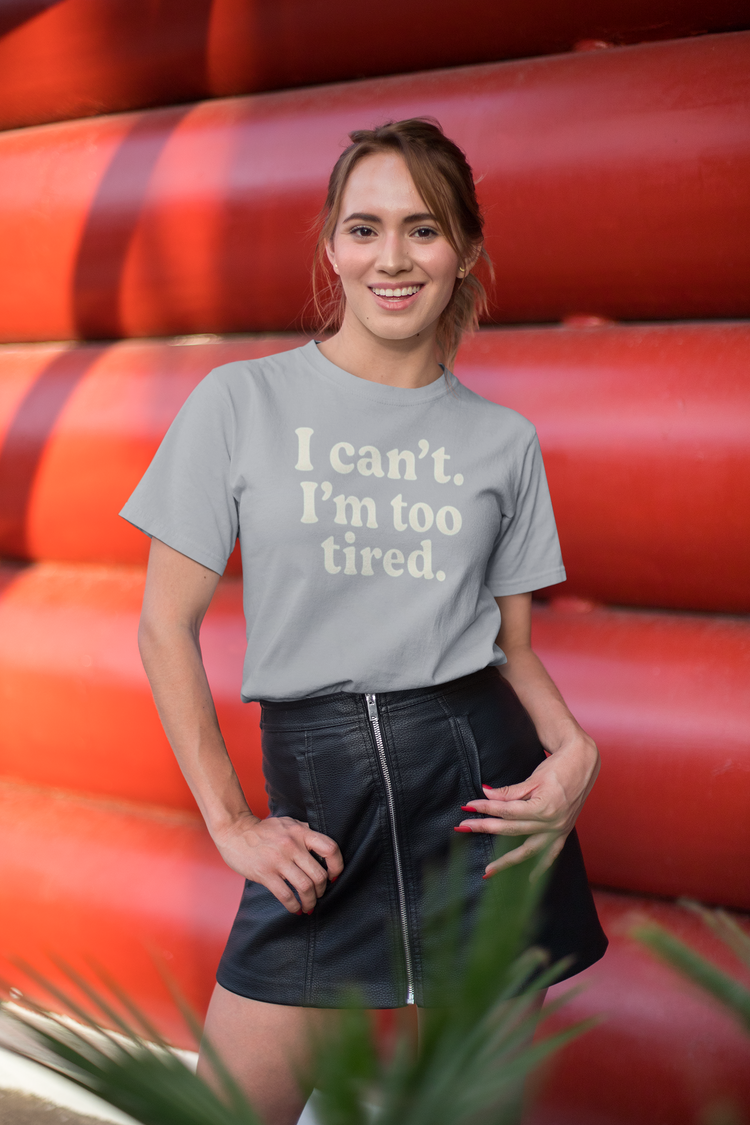 Woman wearing a gray t-shirt with 'I can't. I'm too tired.' text, standing in front of red and orange cylindrical objects.